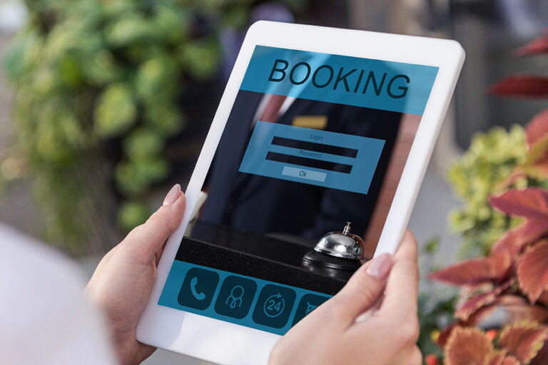 A person holding a tablet displaying an online booking interface with fields for login and password, alongside a service bell on a counter and greenery in the background.