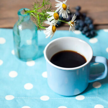 A light blue mug filled with black coffee sits on a polka-dotted blue napkin, accompanied by a small glass vase holding white daisies and green foliage, with some blueberries in the background.