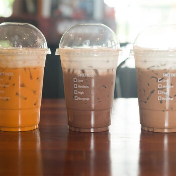 Three clear plastic cups are filled with iced beverages on a wooden table. The left cup contains a yellow-orange drink, the middle cup has a light brown drink with foam, and the right cup holds a darker brown drink. Each cup has a checklist for sweetness levels printed on the side.