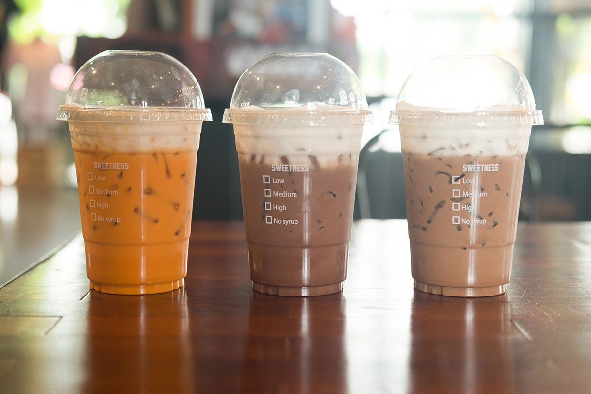 Three clear plastic cups are filled with iced beverages on a wooden table. The left cup contains a yellow-orange drink, the middle cup has a light brown drink with foam, and the right cup holds a darker brown drink. Each cup has a checklist for sweetness levels printed on the side.