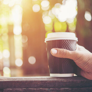 A hand holding a to-go coffee cup with a wavy design, set against a blurred, sunlit outdoor background.