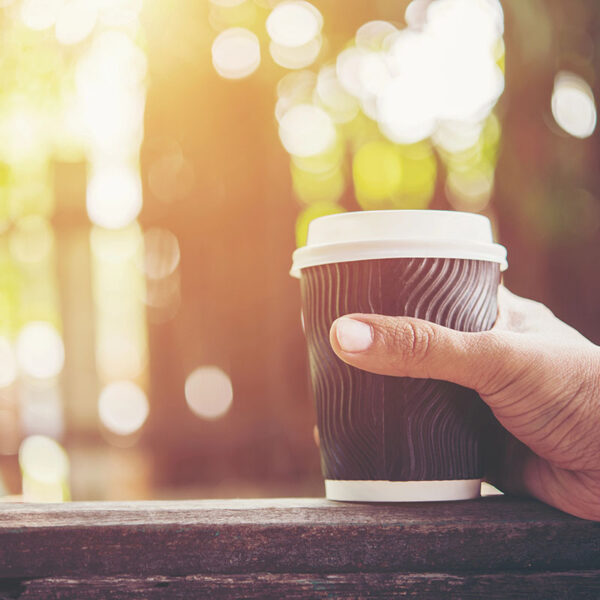 A hand holding a to-go coffee cup with a wavy design, set against a blurred, sunlit outdoor background.