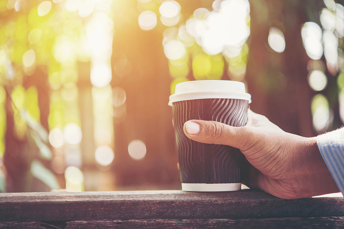 A hand holding a to-go coffee cup with a wavy design, set against a blurred, sunlit outdoor background.