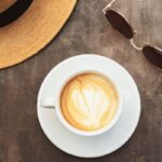 A white cup of latte with heart-shaped foam art sits on a saucer on a wooden table, accompanied by a straw hat and brown sunglasses.