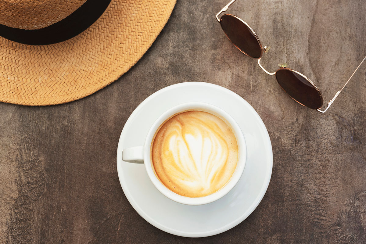 A white cup of latte with heart-shaped foam art sits on a saucer on a wooden table, accompanied by a straw hat and brown sunglasses.