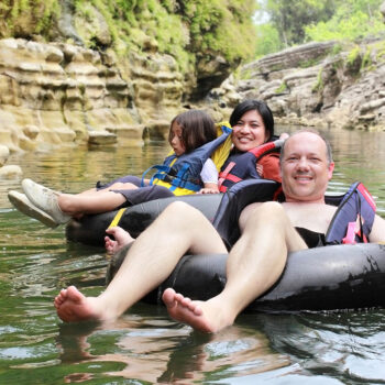 Three people are floating on inner tubes in a calm river surrounded by rocky cliffs and greenery. A child sits in front, while a smiling woman and man relax in the tubes, enjoying the sunny day.