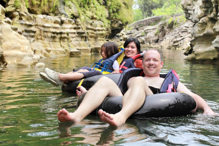 Three people are floating on inner tubes in a calm river surrounded by rocky cliffs and greenery. A child sits in front, while a smiling woman and man relax in the tubes, enjoying the sunny day.
