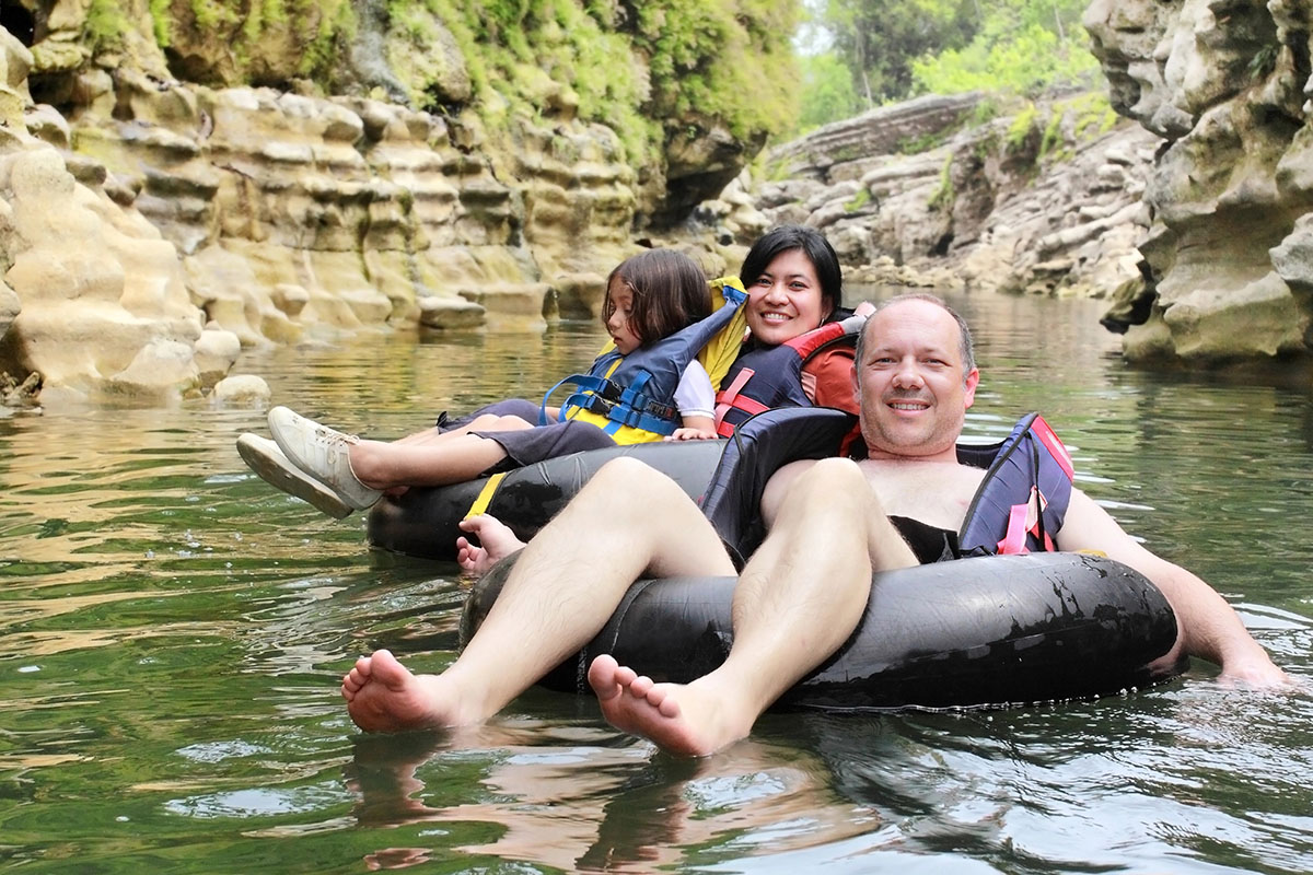 Three people are floating on inner tubes in a calm river surrounded by rocky cliffs and greenery. A child sits in front, while a smiling woman and man relax in the tubes, enjoying the sunny day.