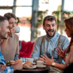 A group of four young adult friends sitting around a small table at a cafe, drinking coffee, eating pastries, and chatting animatedly.