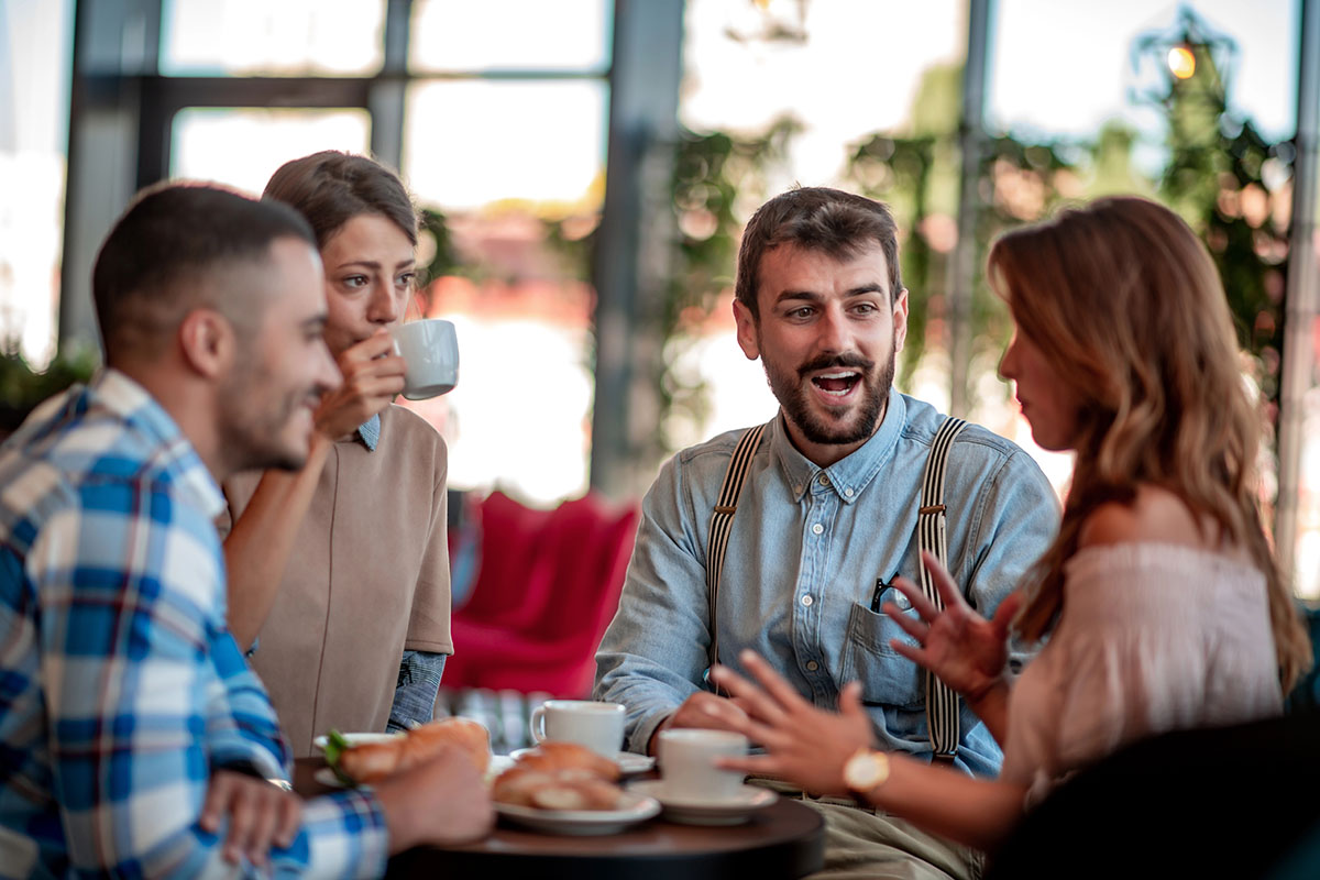 A group of four young adult friends sitting around a small table at a cafe, drinking coffee, eating pastries, and chatting animatedly.