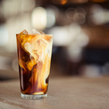A tall glass of iced coffee filled with ice cubes, with milk swirling into the dark brew against a softly blurred cafe background.