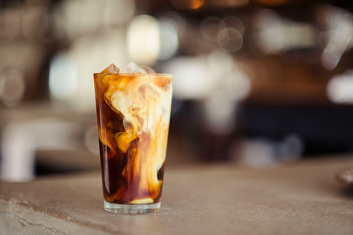 A tall glass of iced coffee filled with ice cubes, with milk swirling into the dark brew against a softly blurred cafe background.