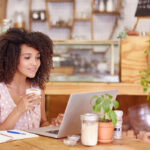 A young woman with curly hair holding a latte while working on a laptop at a café table.