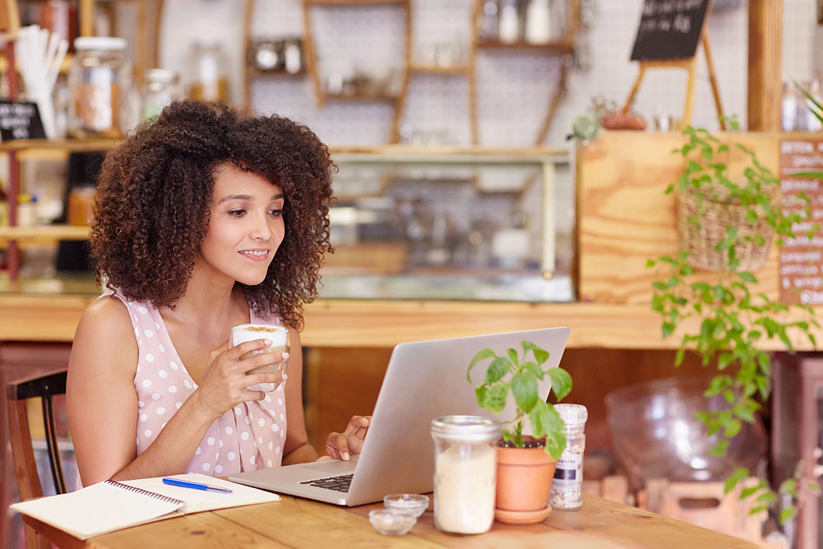 A young woman with curly hair holding a latte while working on a laptop at a café table.