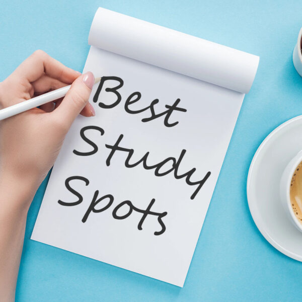 A top-down view of a hand writing "Best Study Spots" on a white notepad, positioned on a light blue desk next to a coffee cup and a small cactus.