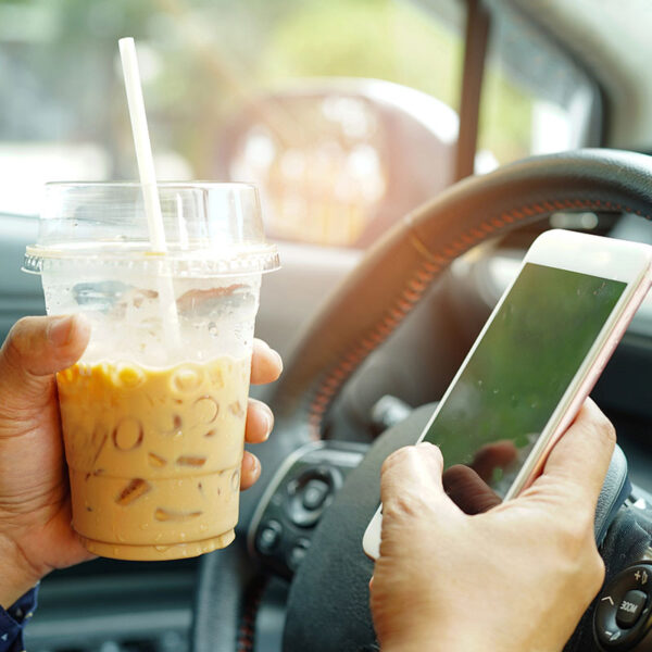 Driver inside a vehicle holding an iced coffee in a plastic cup in one hand and operating a smartphone near the steering wheel with the other.