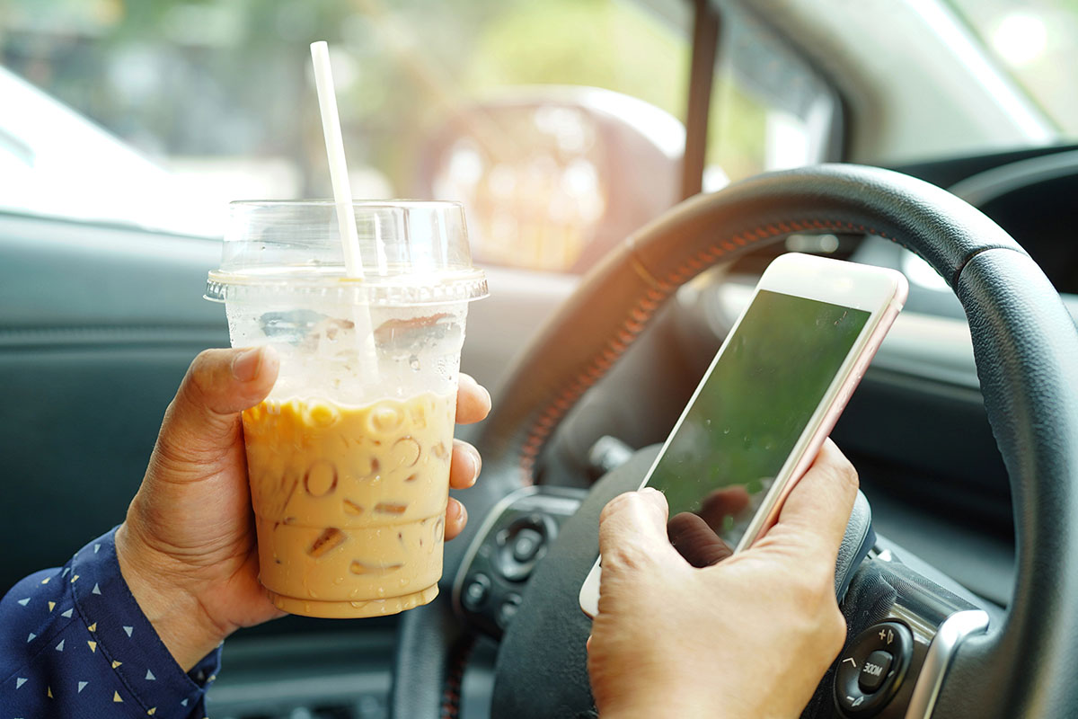 Driver inside a vehicle holding an iced coffee in a plastic cup in one hand and operating a smartphone near the steering wheel with the other.